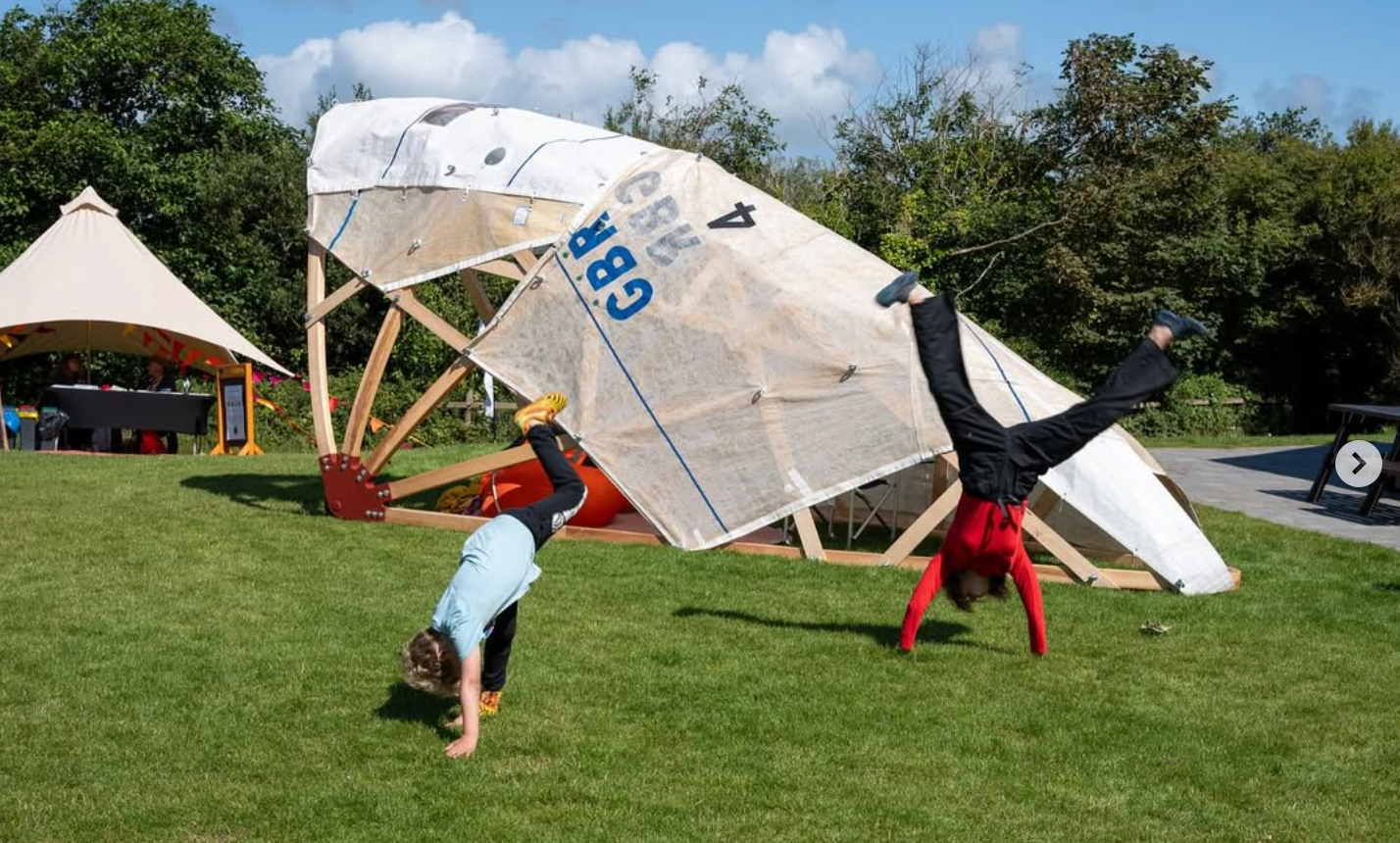 Children play in front of a wooden structure skinned with a ship sale. It's a bright sunny day and the image is joyful.