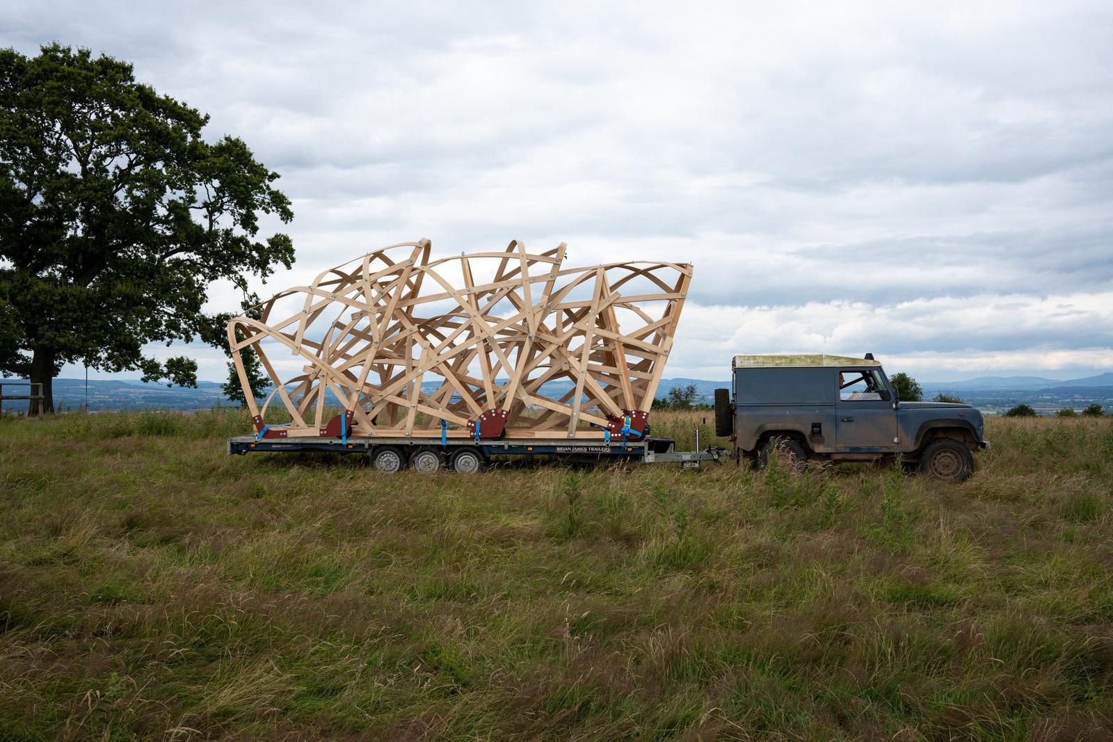 A blue Landrover Defender with a trailer on which sits an architecturally interesting set of structures made of bent wood.
