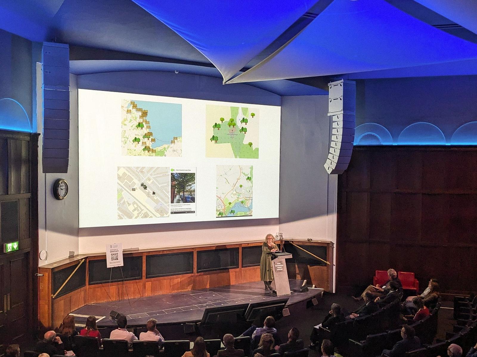 Professor Flora Samuel stands at a lectern in front of an audience with a slide showing maps projected on a screen behind her.